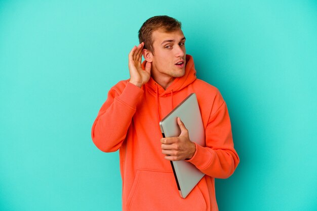 Hombre caucásico joven estudiante sosteniendo un portátil aislado sobre fondo azul tratando de escuchar un chisme.