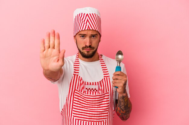 Hombre caucásico joven del fabricante de hielo con los tatuajes que sostienen la cuchara aislada en el fondo rosado que se coloca con la mano extendida que muestra la señal de pare, previniéndole.