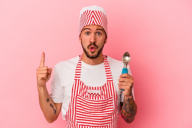 Hombre caucásico joven del fabricante de hielo con los tatuajes que sostienen la cuchara aislada en el fondo rosado que tiene una gran idea, concepto de la creatividad.