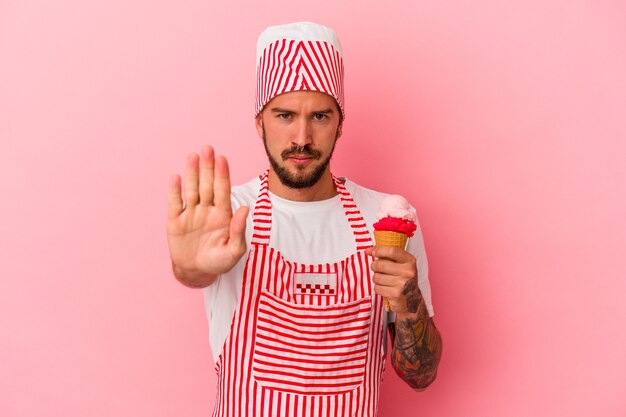 Hombre caucásico joven del fabricante de hielo con tatuajes que sostienen el helado aislado en el fondo rosado que se coloca con la mano extendida que muestra la señal de stop, que le impide.