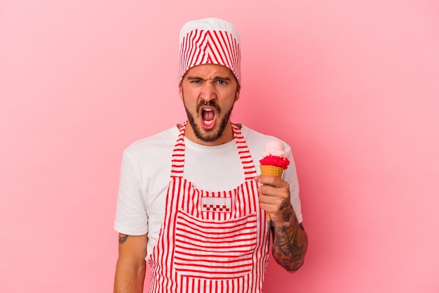 Hombre caucásico joven fabricante de hielo con tatuajes sosteniendo helado aislado sobre fondo rosa gritando muy enojado y agresivo.