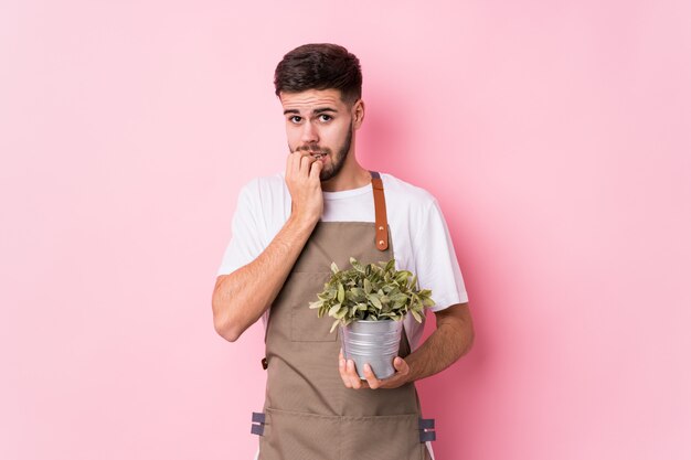 El hombre caucásico joven del jardinero que sostenía una planta aisló morderse las uñas, nervioso y muy ansioso.