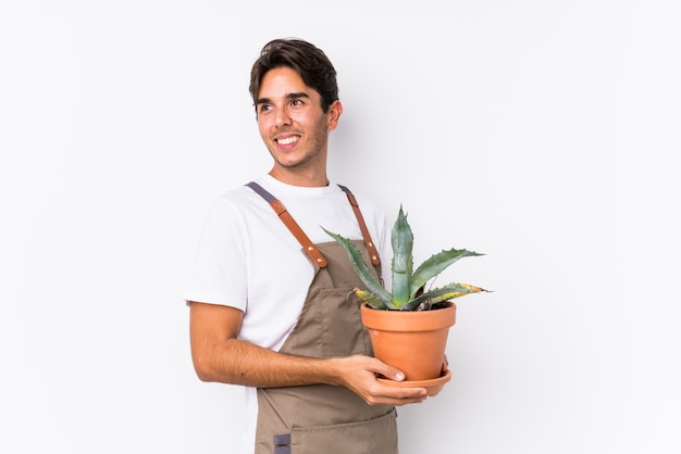 El hombre caucásico joven del jardinero que sostiene una planta aislada mira a un lado sonriente, alegre y agradable.