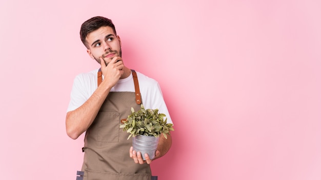 Hombre caucásico joven jardinero sosteniendo una planta aislada mirando hacia los lados con expresión dudosa y escéptica.