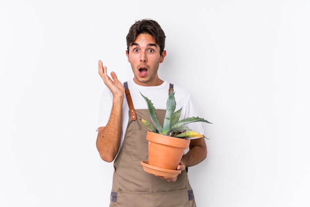 Hombre caucásico joven jardinero sosteniendo una planta aislada sorprendido y conmocionado.