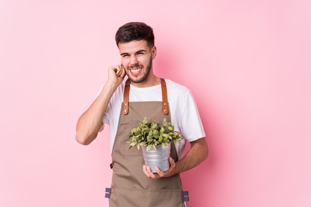 Hombre caucásico joven jardinero sosteniendo una planta cubriendo las orejas con las manos.