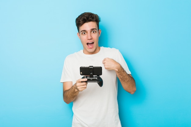 Hombre caucásico joven jugando videojuegos con teléfono sorprendido apuntando a sí mismo, sonriendo ampliamente.