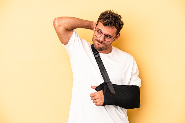 Foto hombre caucásico joven con la mano rota aislada sobre fondo amarillo tocando la parte posterior de la cabeza, pensando y haciendo una elección.