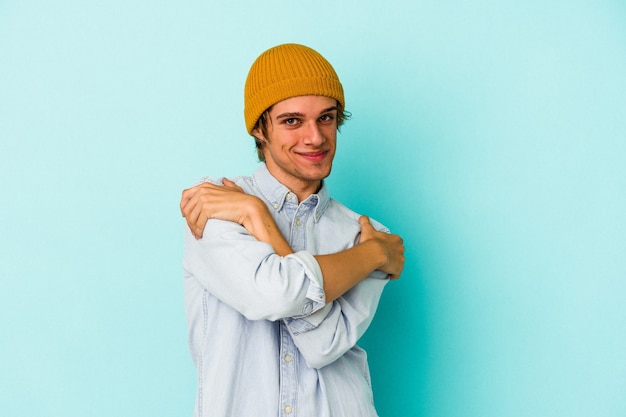 Hombre caucásico joven con maquillaje aislado sobre fondo azul abrazos, sonriendo despreocupado y feliz.