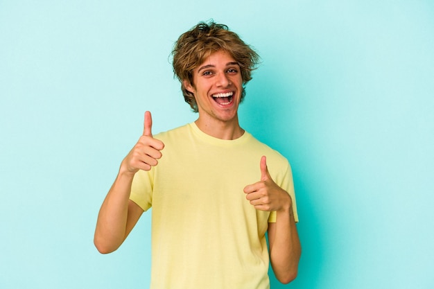 Hombre caucásico joven con maquillaje aislado sobre fondo azul levantando ambos pulgares, sonriente y confiado.