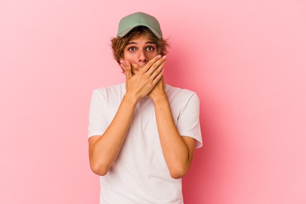 Hombre caucásico joven con maquillaje aislado sobre fondo rosa cubriendo la boca con las manos mirando preocupado.