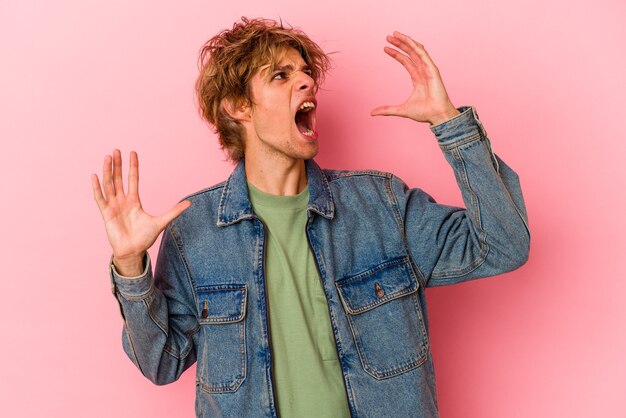 Hombre caucásico joven con maquillaje aislado sobre fondo rosa gritando al cielo, mirando hacia arriba, frustrado.