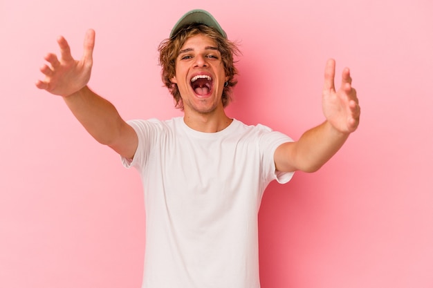 Hombre caucásico joven con maquillaje aislado sobre fondo rosa se siente confiado dando un abrazo a la cámara.