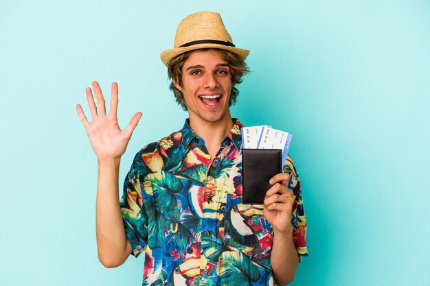 Hombre caucásico joven con maquillaje con pasaporte aislado sobre fondo azul sonriendo alegre mostrando el número cinco con los dedos.