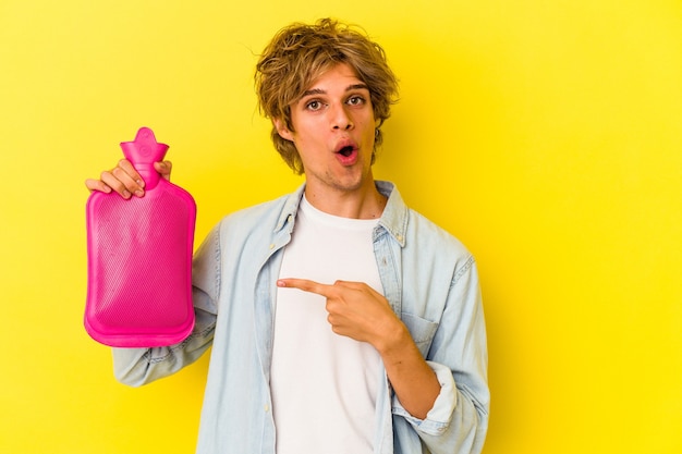 Hombre caucásico joven con maquillaje sosteniendo una bolsa de agua caliente aislada sobre fondo amarillo apuntando hacia el lado