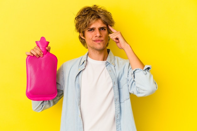 Hombre caucásico joven con maquillaje sosteniendo una bolsa de agua caliente aislada sobre fondo amarillo señalando la sien con el dedo, pensando, centrado en una tarea.