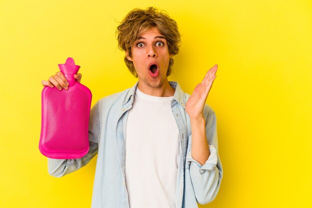 Hombre caucásico joven con maquillaje sosteniendo una bolsa de agua caliente aislada sobre fondo amarillo sorprendido y consternado.