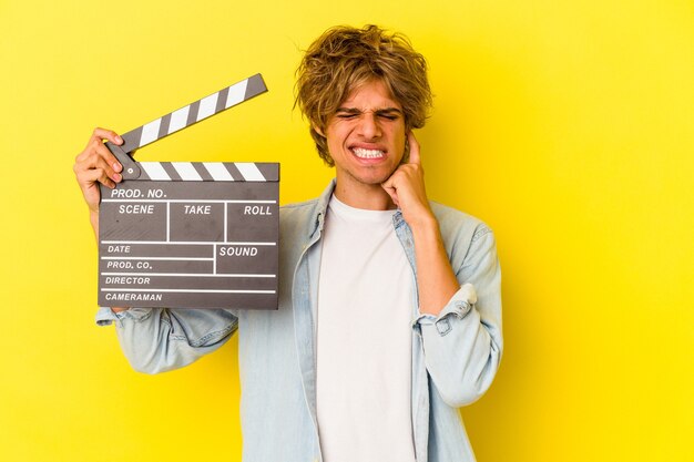 Hombre caucásico joven con maquillaje sosteniendo claqueta aislado sobre fondo amarillo cubriendo las orejas con las manos.