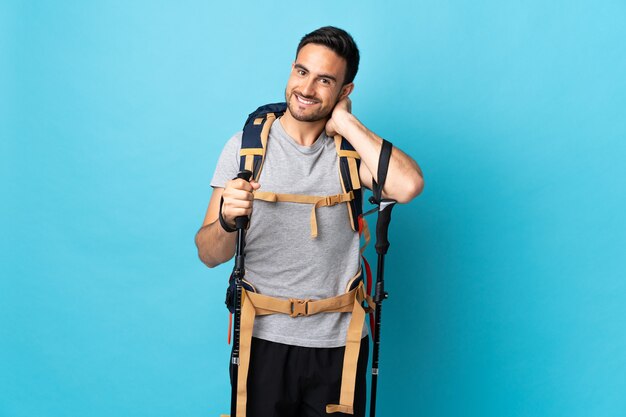 Hombre caucásico joven con mochila y bastones de trekking aislado en la pared azul riendo