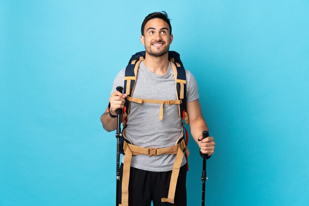 Hombre caucásico joven con mochila y bastones de trekking aislado sobre fondo azul mirando hacia arriba mientras sonríe