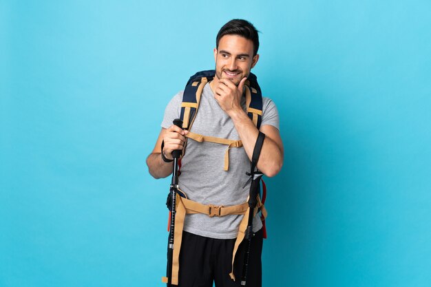 Hombre caucásico joven con mochila y bastones de trekking aislados en azul mirando hacia el lado y sonriendo