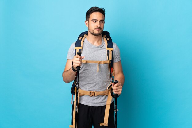 Hombre caucásico joven con mochila y bastones de trekking aislados en azul que tiene dudas mientras mira de lado