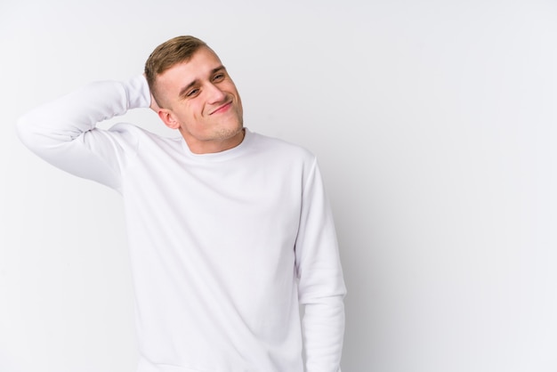 Hombre caucásico joven en la pared blanca tocando la parte posterior de la cabeza, pensando y haciendo una elección.