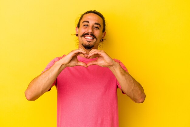 Hombre caucásico joven con pelo largo aislado sobre fondo amarillo sonriendo y mostrando una forma de corazón con las manos.
