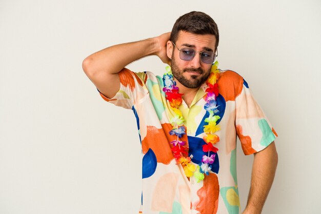 Hombre caucásico joven que baila en una fiesta hawaiana aislada en la pared blanca que toca la parte posterior de la cabeza, pensando y haciendo una elección.