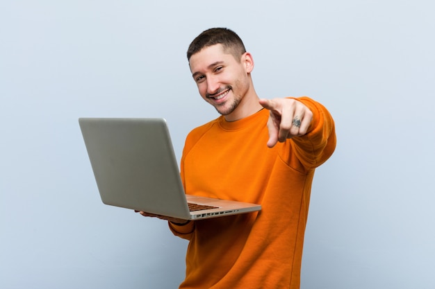 Hombre caucásico joven que celebra sonrisas alegres de una computadora portátil que señalan al frente.