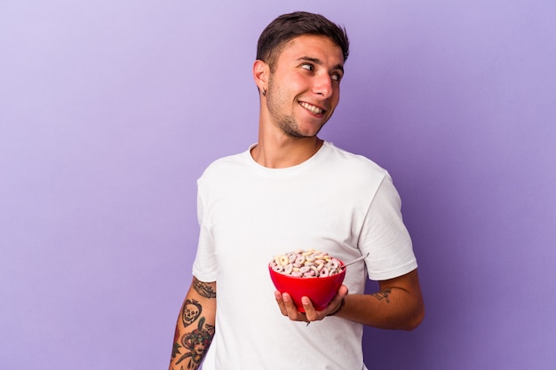 El hombre caucásico joven que come los cereales aislado en el fondo púrpura mira a un lado sonriente, alegre y agradable.