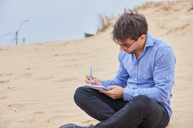 Hombre caucásico joven que escribe una carta o poesía en su cuaderno, sentado en la arena de la playa, en traje formal.