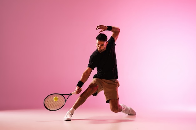 Hombre caucásico joven que juega al tenis aislado en la acción de la pared rosada y el concepto del movimiento