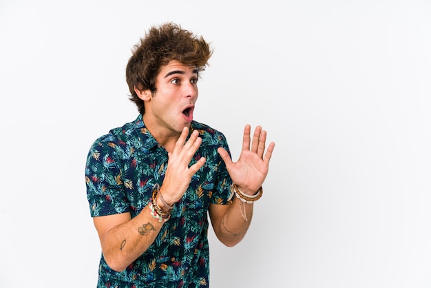 El hombre caucásico joven que lleva una camiseta de la flor aislada grita ruidosamente, mantiene los ojos abiertos y las manos tensas.