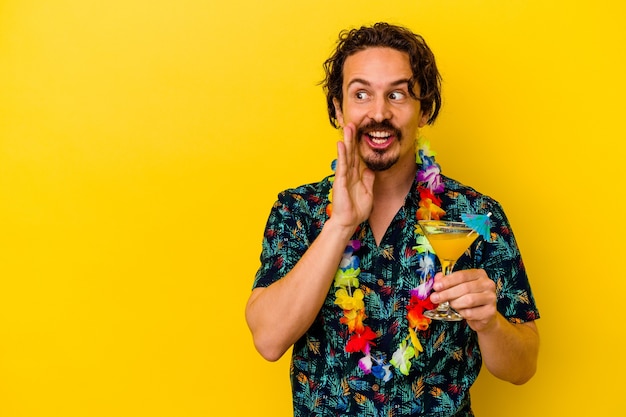 Hombre caucásico joven que lleva un collar hawaiano sosteniendo un cóctel aislado sobre fondo amarillo está diciendo una noticia secreta de frenado caliente y mirando a un lado