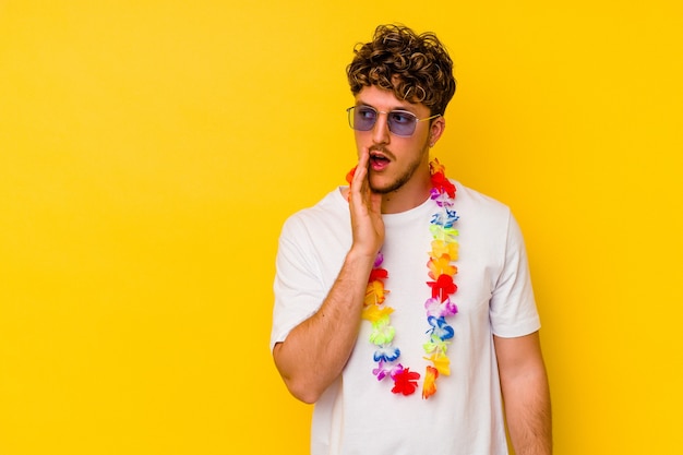 Hombre caucásico joven que lleva un material de fiesta hawaiano aislado en la pared amarilla está diciendo una noticia secreta de frenado caliente y mirando a un lado