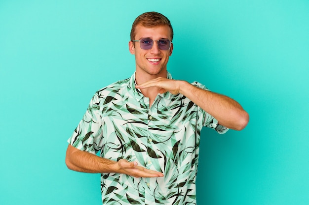 Hombre caucásico joven que lleva una ropa de verano aislada en azul que sostiene algo con ambas manos, presentación del producto.