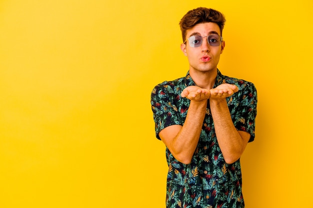 Hombre caucásico joven que llevaba una camisa hawaiana aislada en la pared amarilla doblando los labios y sosteniendo las palmas para enviar un beso de aire.