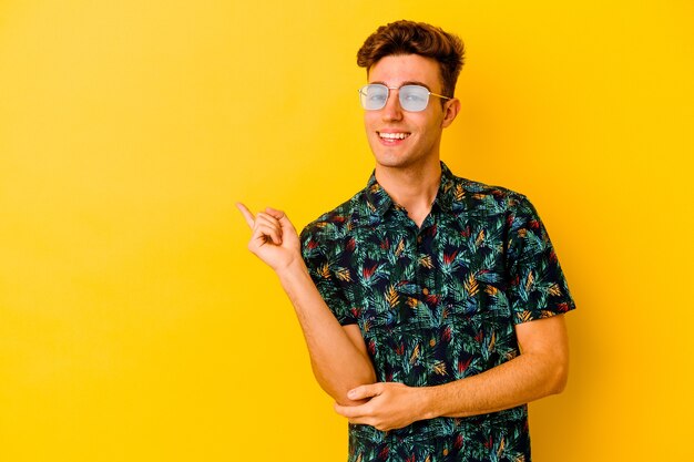 Hombre caucásico joven que llevaba una camisa hawaiana aislado en la pared amarilla sonriendo alegremente apuntando con el dedo índice lejos