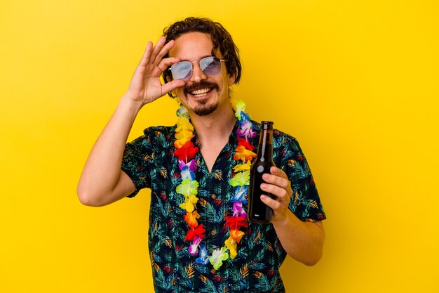 Hombre caucásico joven que llevaba un collar hawaiano sosteniendo una cerveza emocionado manteniendo el gesto ok en el ojo.