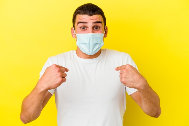 Hombre caucásico joven que llevaba una protección para el coronavirus aislado en la pared amarilla sorprendido señalando con el dedo, sonriendo ampliamente.