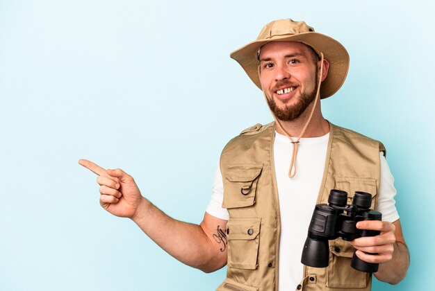 Hombre caucásico joven que mira a los animales a través de binoculares aislados sobre fondo azul sonriendo y apuntando a un lado, mostrando algo en el espacio en blanco.