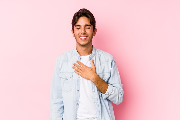 Hombre caucásico joven que presenta en una pared rosada aislada se ríe a carcajadas manteniendo la mano en el pecho.