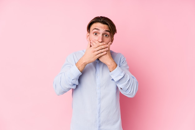 Hombre caucásico joven que presenta en una pared rosada aislada sorprendida cubriendo la boca con las manos.