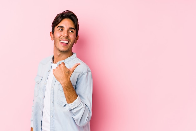 El hombre caucásico joven que presenta en una pared rosada señala con el dedo pulgar lejos, riendo y despreocupado.