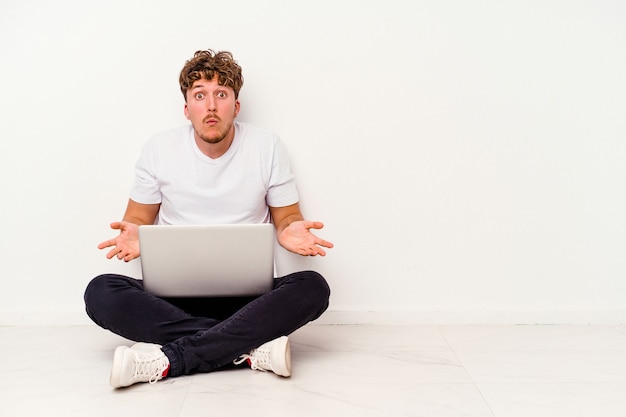 Hombre caucásico joven que se sienta en el suelo que sostiene en la computadora portátil aislada en el fondo blanco encoge de hombros y abre los ojos confundidos.