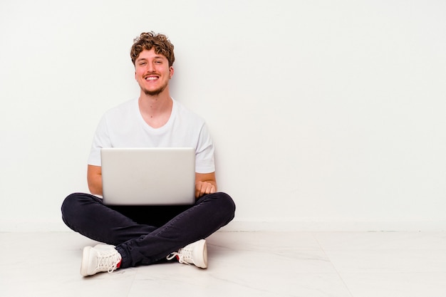 Hombre caucásico joven que se sienta en el suelo que sostiene en la computadora portátil aislada en el fondo blanco feliz, sonriente y alegre.