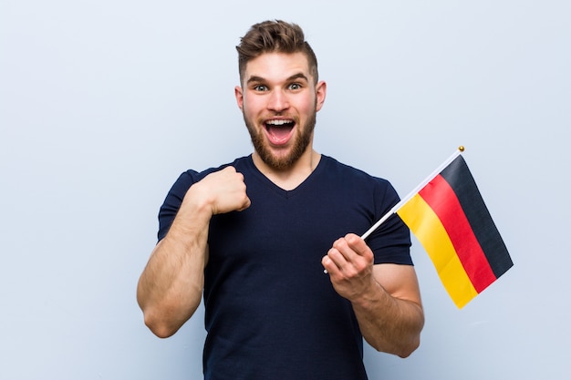 El hombre caucásico joven que sostenía una bandera de Alemania sorprendió señalar a sí mismo, sonriendo ampliamente.