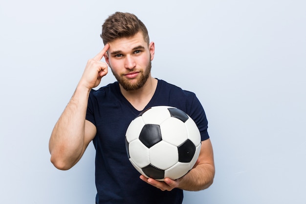 Hombre caucásico joven que sostiene un balón de fútbol apuntando su sien con el dedo, pensando, centrado en una tarea.