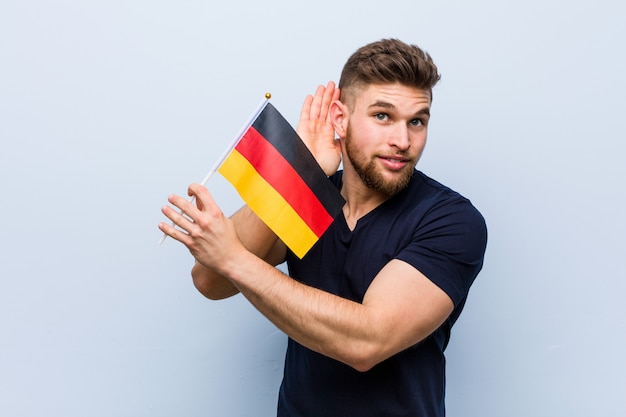 Hombre caucásico joven que sostiene una bandera de Alemania que intenta escuchar un chisme.